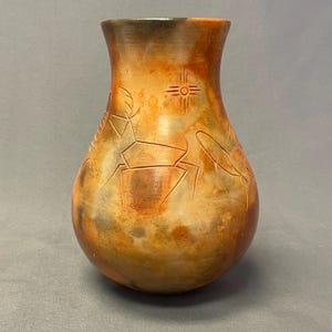 May include: A rounded ceramic vase with a mottled orange, brown, and black finish. The surface features etched line drawings of a four-legged animal and a sun symbol. The vase has a wide, curved body that tapers toward a smooth, open rim.