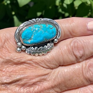 May include: A silver ring with a turquoise stone set in a detailed design. The ring is on a person's finger.