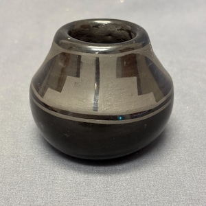 May include: A small, round, black ceramic pot with a wide opening. The pot features a geometric design in gray and black, with a band of black at the base. The design is symmetrical and appears to be hand-painted.