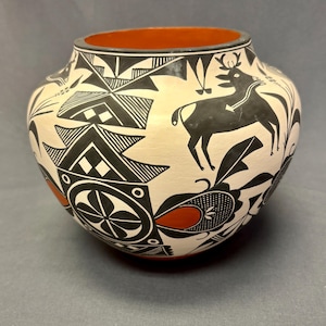 May include: Hand-painted ceramic pot with a cream-colored background and black and red geometric and animal designs. The pot features a deer silhouette and a floral pattern. The rim is black and the interior is orange.