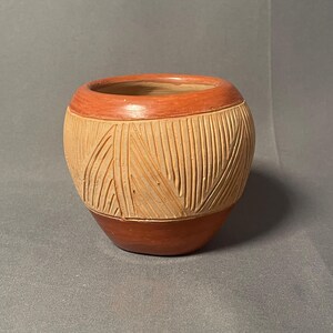 May include: A handcrafted, terracotta-colored ceramic pot with a textured, carved design. The pot features a reddish-brown rim and base, with a lighter tan body adorned with vertical and angled lines. The vessel has a rounded shape.