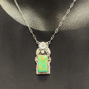 May include: A silver pendant necklace featuring a rectangular turquoise stone with gold veining. The pendant is framed by ornate silver detailing, including leaves and a floral design at the top. The necklace has a delicate silver chain.