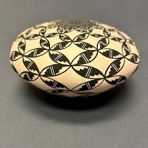 May include: A hand-painted, oval-shaped ceramic piece with a cream-colored base. The surface is adorned with a repeating black geometric pattern of interlocking shapes. The design covers the entire top surface of the object.