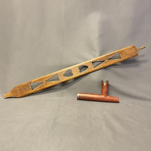 May include: A long, narrow, light brown wooden slat featuring cut-out geometric shapes including triangles and two central heart shapes, accompanied by a separate, smaller, smooth reddish-brown wooden T-shaped pipe with a metallic rim at the top.