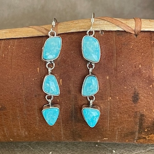 May include: A pair of dangle earrings with three turquoise stones each. The stones are a light blue color and are set in silver.