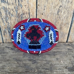 May include: A blue, red, white, and black beaded oval with a geometric design. The beadwork is intricate and detailed.