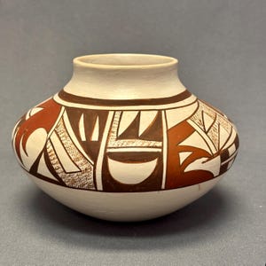 May include: A handcrafted pottery vessel with a rounded body and a short neck. The off-white surface is adorned with geometric patterns in brown and reddish-brown, creating a striking visual contrast. The design includes triangles, lines, and curved shapes.