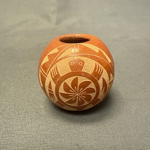 May include: A small, round, orange-brown pottery vase with a carved turtle design. The turtle has a flower-like pattern on its shell. The vase has a small opening at the top and is likely handmade.