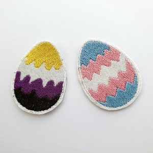 May include: Two embroidered iron-on patches shaped like eggs. One is yellow, white, purple, and black, and the other is blue, pink, and white.