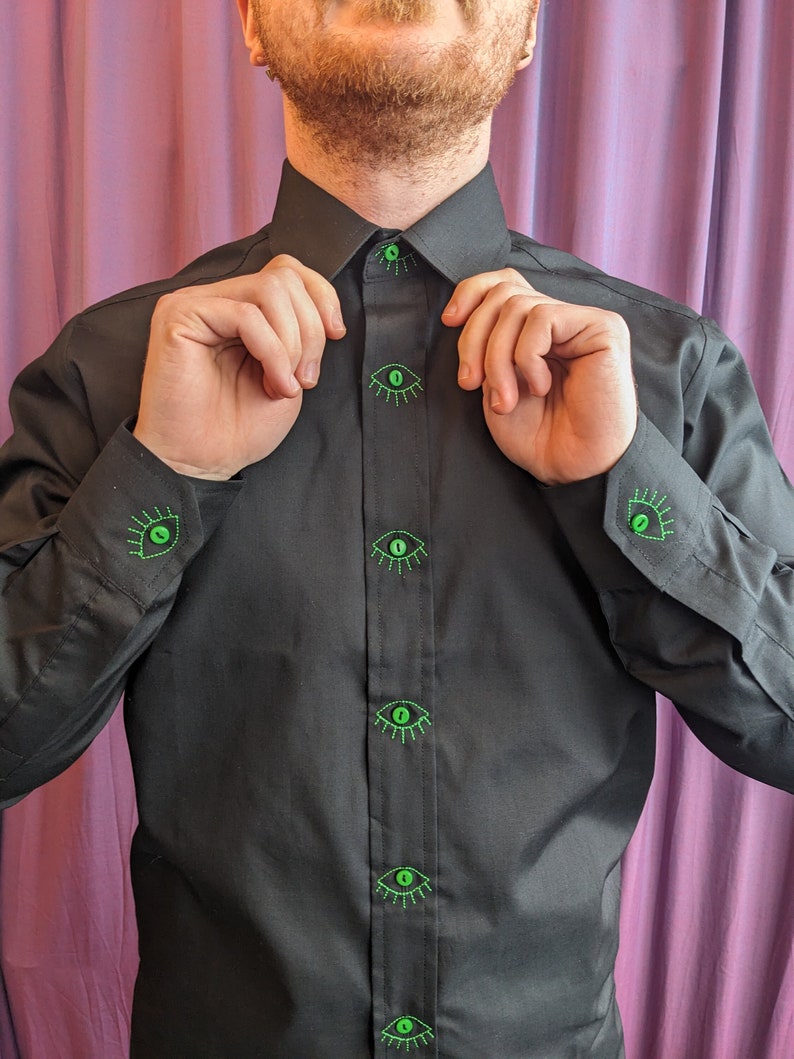 May include: A black dress shirt with green embroidered eyes on the front and cuffs. The shirt has a button-up collar and long sleeves.