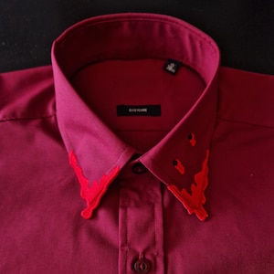 May include: A burgundy button-down shirt with a red embroidered collar. The collar has a jagged, dripping design.