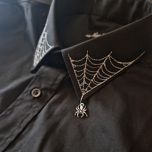 Może przedstawiać: Czarna koszula zapinana na guziki z haftowanym wzorem białej pajęczyny na kołnierzyku. Srebrny wisiorek w kształcie pająka zwisa z dolnej części pajęczyny.