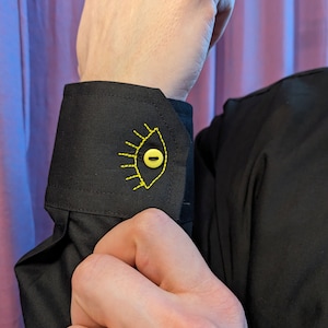 May include: Black shirt cuff with a yellow eye embroidered on it. The eye has a yellow button in the center.