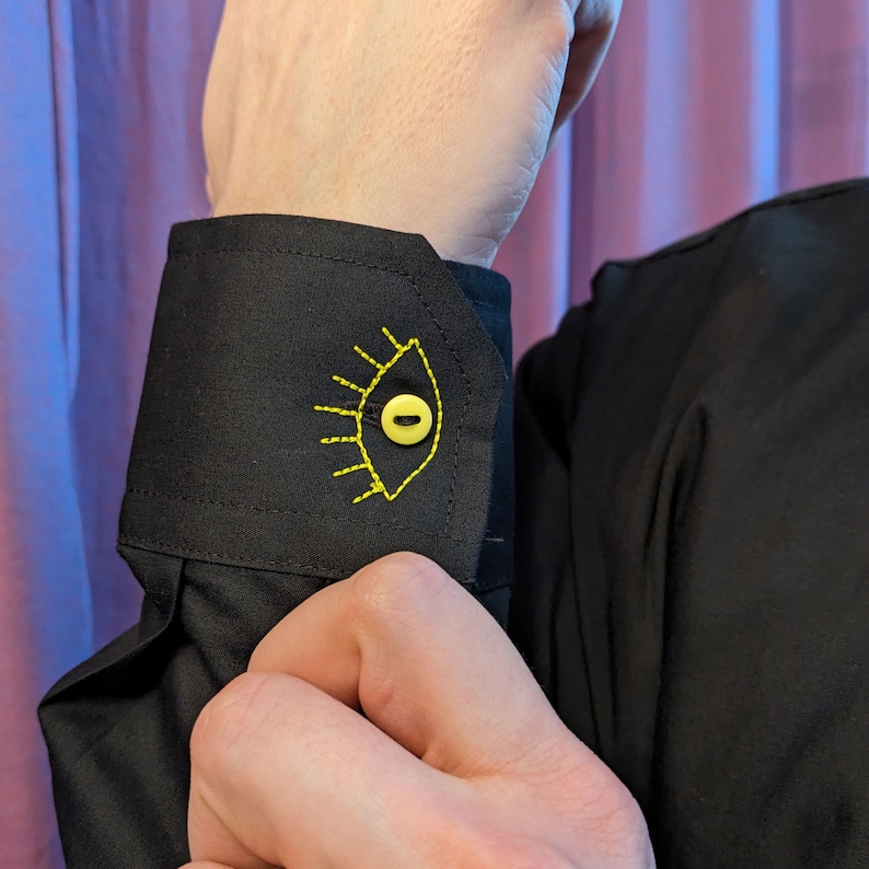 May include: Black shirt cuff with a yellow eye embroidered on it. The eye has a yellow button in the center.