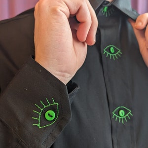 May include: A black button-down shirt with green eye-shaped buttons. The buttons have green stitching around the edges, resembling eyelashes.