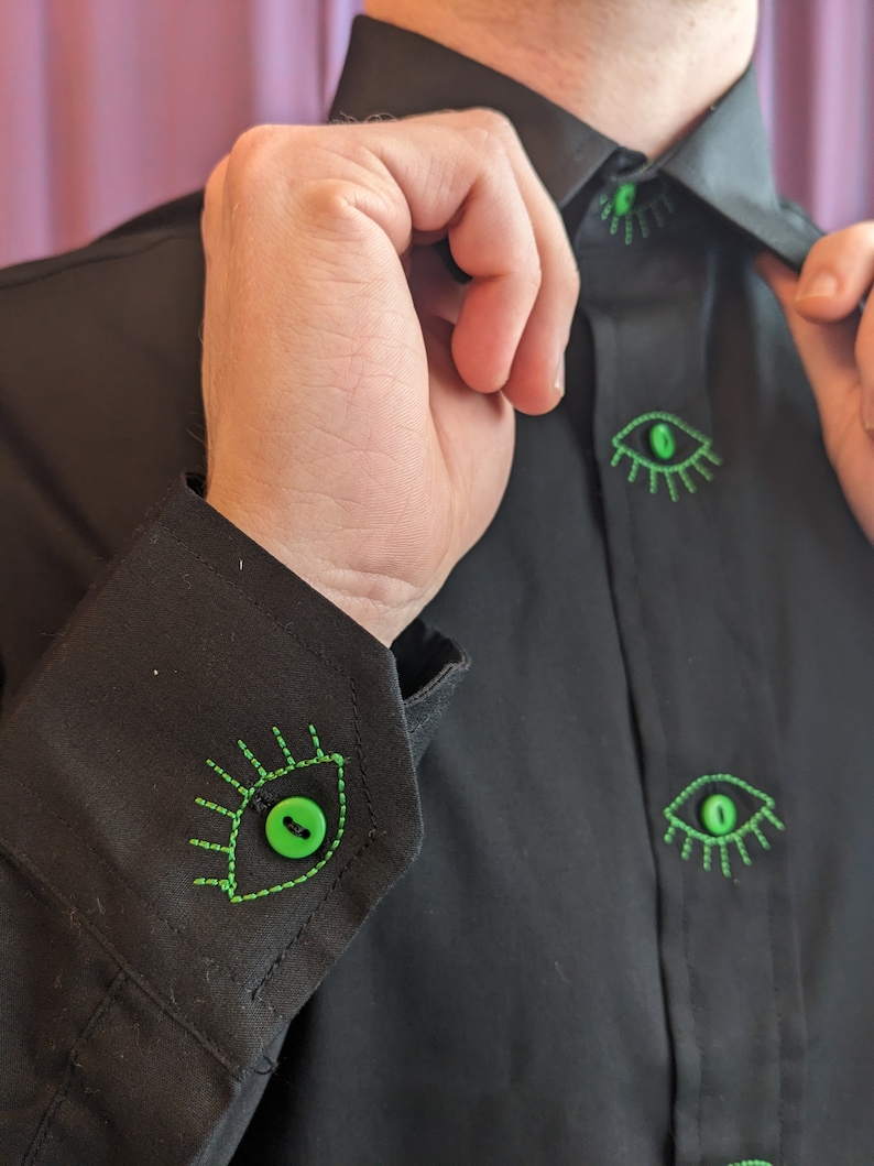 May include: A black button-down shirt with green eye-shaped buttons. The buttons have green stitching around the edges, resembling eyelashes.