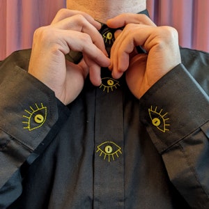 May include: A black button-down shirt with yellow embroidered eyes on the cuffs and front. The eyes have a yellow outline and a black center.