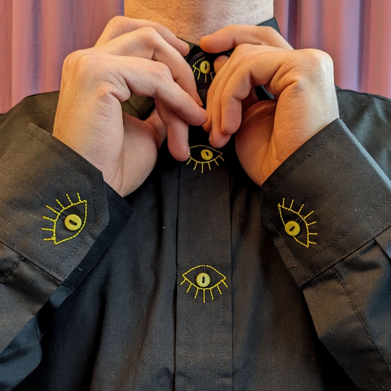 May include: A black button-down shirt with yellow embroidered eyes on the cuffs and front. The eyes have a yellow outline and a black center.