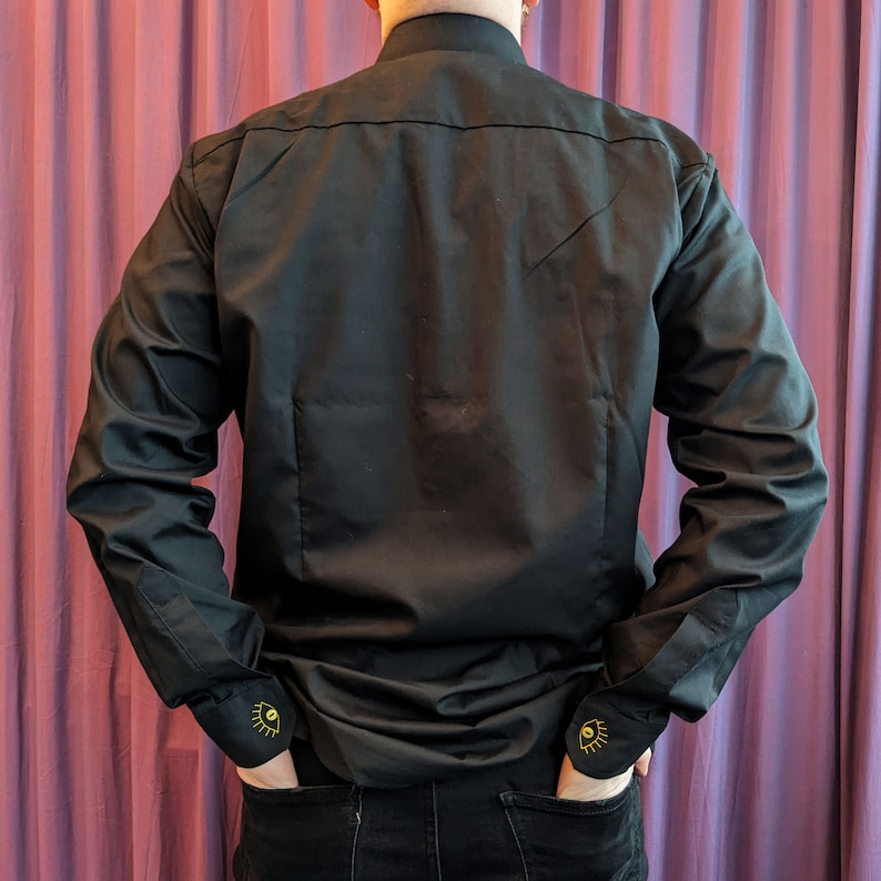 May include: A black long-sleeve shirt with a mock neck collar and gold eye embroidery on the cuffs.