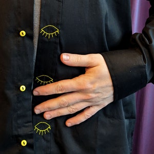 May include: A black button-down shirt with yellow embroidered eyes on the front. The shirt is worn by a person with their hand resting on their stomach.