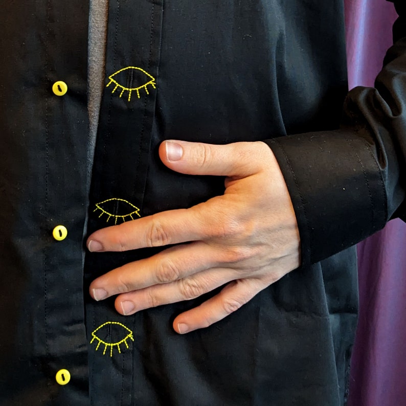 May include: A black button-down shirt with yellow embroidered eyes on the front. The shirt is worn by a person with their hand resting on their stomach.