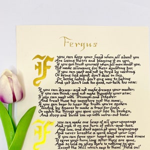 May include: A cream-colored card featuring the poem If by Rudyard Kipling written in black calligraphy with gold illuminated letters. A single artificial tulip with a purple and white flower and green stem sits on the left, and a small turquoise stone is on the bottom right.