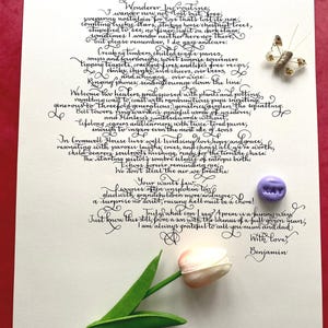 May include: A white sheet of paper featuring black calligraphy text titled For Mum and Dad, signed by Benjamin. The paper is decorated with a small, glittery butterfly ornament, a round purple bead with the word KAY, and a single pink tulip flower.