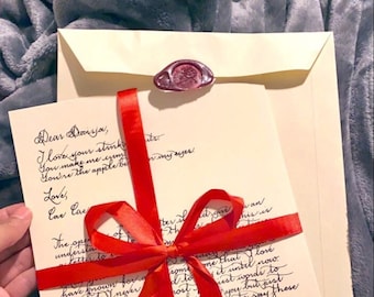 May include: A cream-coloured letter with handwritten black cursive text, tied with a bright red satin ribbon bow. The letter is tucked into a matching cream envelope sealed with a dark red wax stamp featuring a rose design.