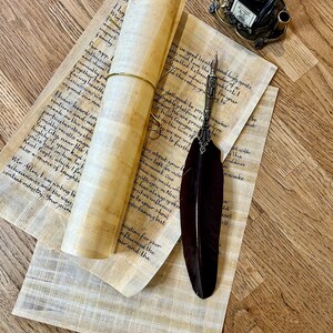 May include: A rolled up scroll of parchment paper with a quill pen and an inkwell on a wooden surface. The scroll is tied with a brown string. The quill pen is black with a silver tip. The inkwell is black with a silver lid and base. The wooden surface is light brown with a grain pattern.