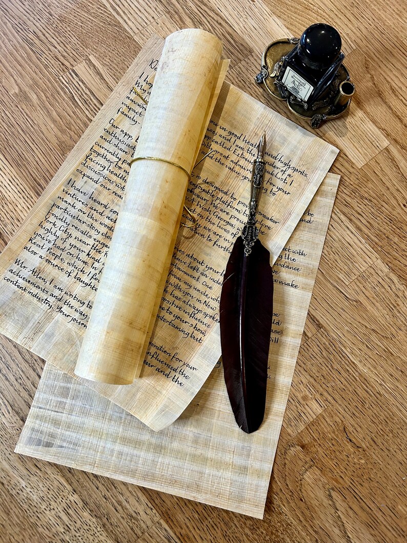 May include: A rolled up scroll of parchment paper with a quill pen and an inkwell on a wooden surface. The scroll is tied with a brown string. The quill pen is black with a silver tip. The inkwell is black with a silver lid and base. The wooden surface is light brown with a grain pattern.