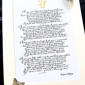 May include: A white paper print featuring the poem If by Rudyard Kipling in black calligraphy, topped with a gold monogram. A small gold butterfly embellishment is attached to the bottom left corner, and a smooth grey stone rests at the top right.