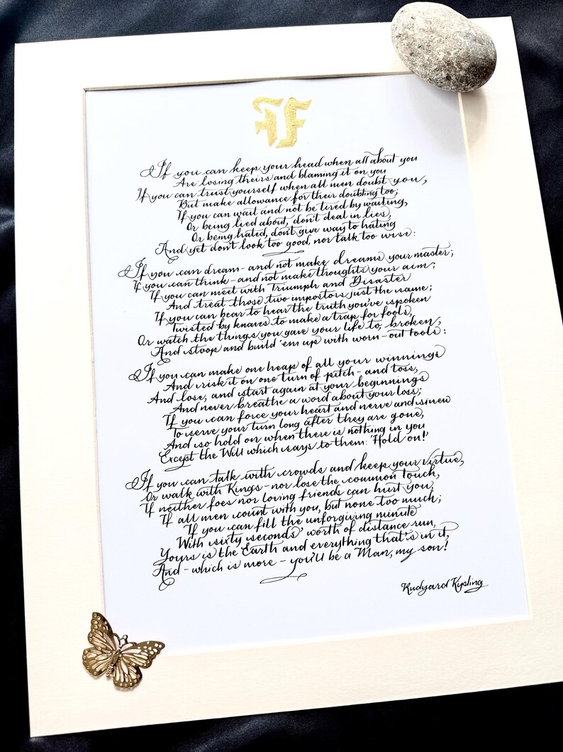 May include: A white paper print featuring the poem If by Rudyard Kipling in black calligraphy, topped with a gold monogram. A small gold butterfly embellishment is attached to the bottom left corner, and a smooth grey stone rests at the top right.