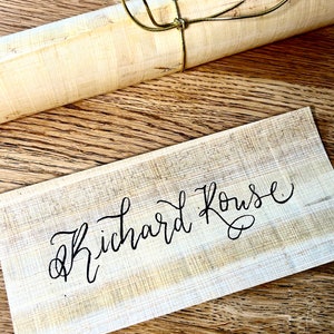 May include: A close-up of a wooden sign with the name "Richard Rouse" written in black calligraphy. The sign is on a wooden surface with a rolled-up piece of paper tied with a gold cord.