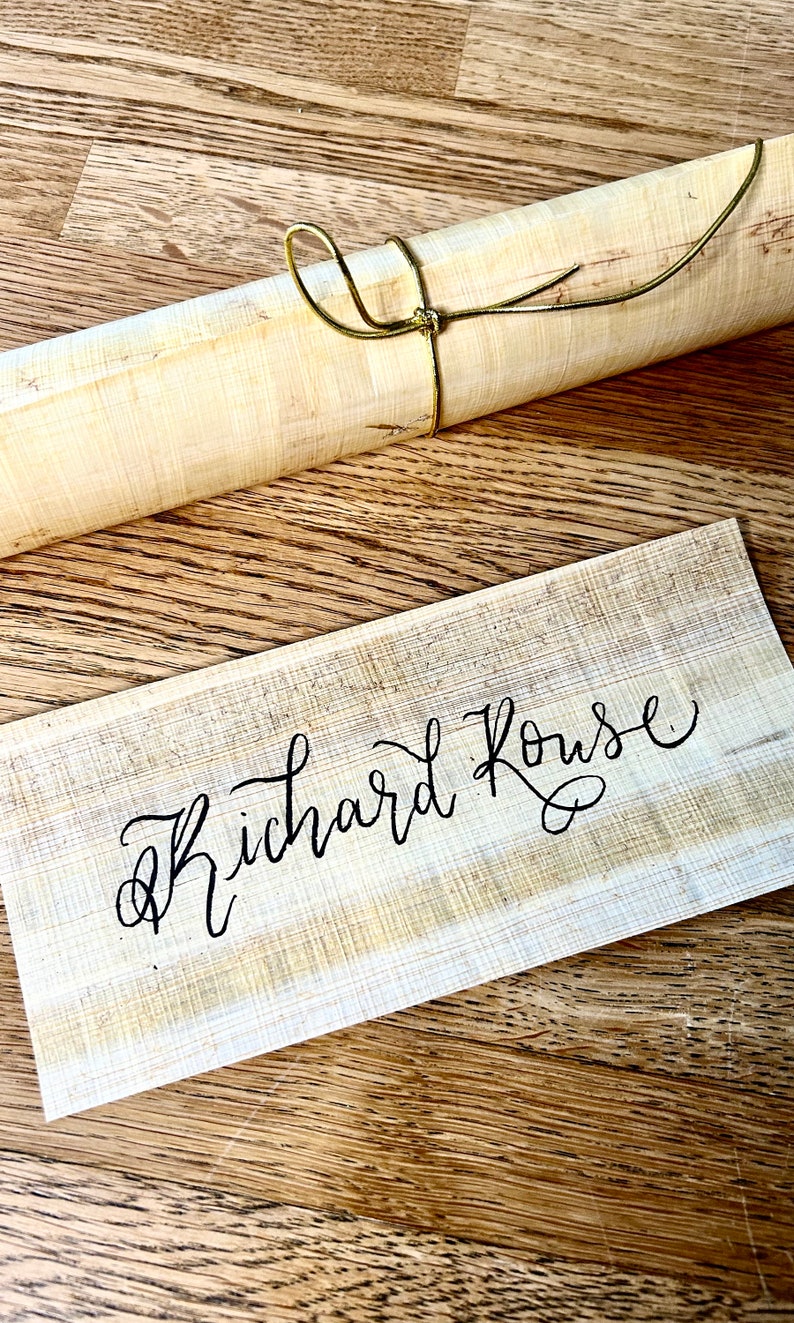 May include: A close-up of a wooden sign with the name "Richard Rouse" written in black calligraphy. The sign is on a wooden surface with a rolled-up piece of paper tied with a gold cord.