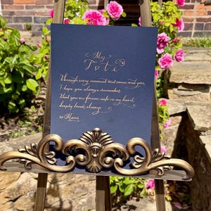 Puede incluir: Un letrero rectangular azul oscuro con texto dorado en cursiva, exhibido en un caballete de metal bronce ornamentado de estilo antiguo. El texto dice: My Tot, Through every moment that comes and goes, I hope you always remember that you are forever safe in my heart, deeply loved, always, xo Mami.