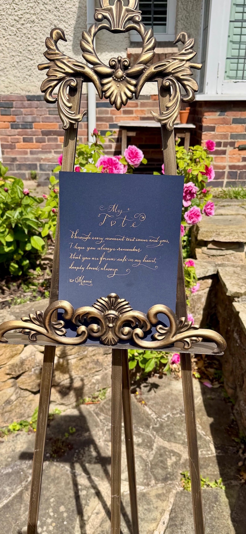 Puede incluir: Un letrero rectangular azul oscuro con texto dorado en cursiva, exhibido en un caballete de metal bronce ornamentado de estilo antiguo. El texto dice: My Tot, Through every moment that comes and goes, I hope you always remember that you are forever safe in my heart, deeply loved, always, xo Mami.