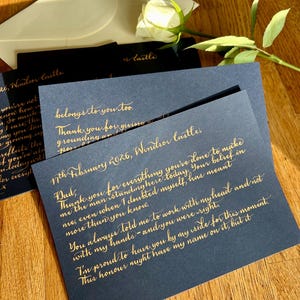 Puede incluir: Una tarjeta azul oscuro con elegante caligraf&iacute;a dorada fechada el 17 de febrero de 2026, Windsor Castle, dirigida a pap&aacute;. La tarjeta se acompa&ntilde;a de un sobre blanco y una rosa blanca, creando un conjunto de papeler&iacute;a formal y sofisticado.