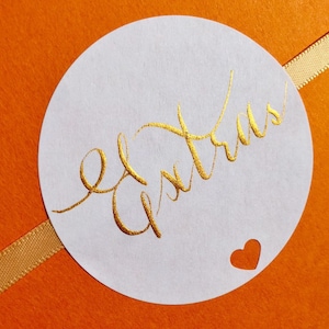 May include: A white round sticker with a gold handwritten inscription that says "El Atrevido" and a small heart cut out in the lower right corner.