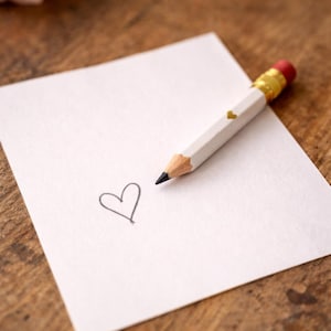Könnte beinhalten: Ein weißer Bleistift mit goldenem Band und rotem Radiergummi liegt auf einem weißen Blatt Papier. Auf dem Papier ist ein kleines, schwarzes Herz gezeichnet. Der Bleistift hat ein kleines goldenes Herz an der Seite. Der Hintergrund ist eine Holzoberfläche.
