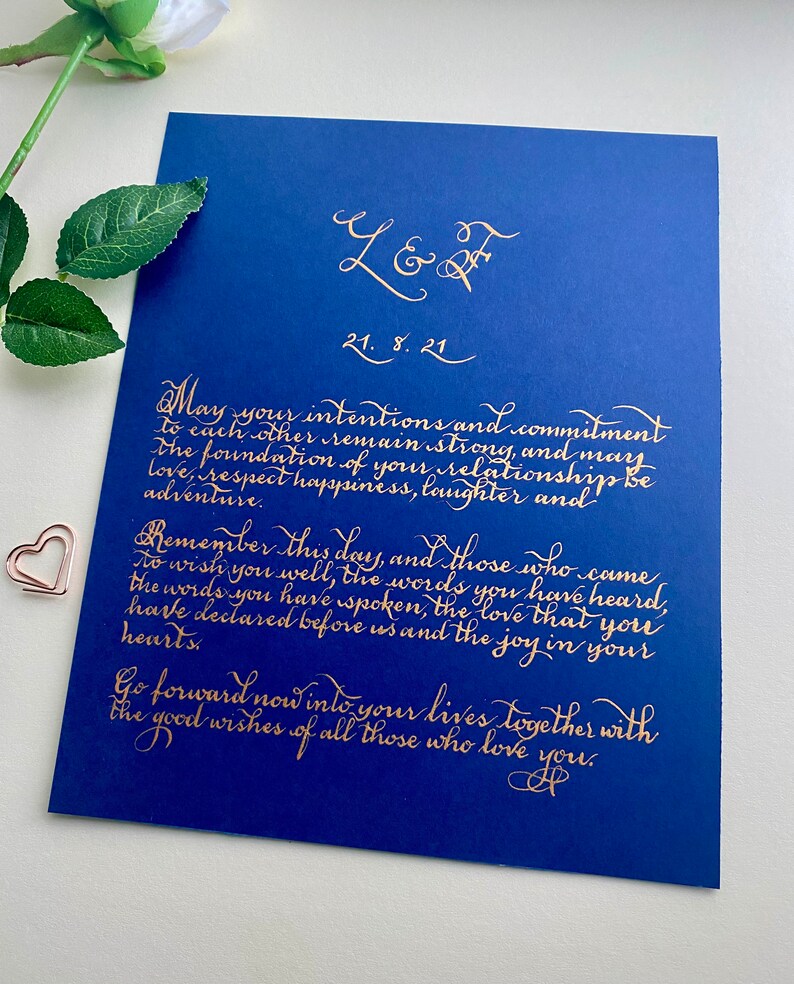 Puede incluir: Una tarjeta rectangular de color azul profundo con elegante caligraf&iacute;a dorada que dice L & F, 21. 8. 21, y un mensaje sincero sobre el compromiso y el amor. Un peque&ntilde;o clip en forma de coraz&oacute;n de oro rosa se encuentra a la izquierda.