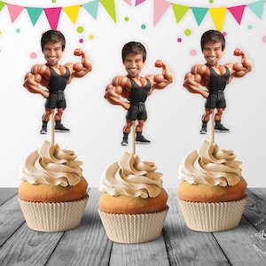 Puede incluir: Tres adornos para cupcakes con un hombre de dibujos animados con un físico musculoso que lleva un tanque negro y pantalones cortos negros. Los adornos están encima de cupcakes con glaseado marrón.