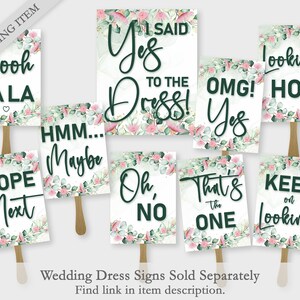 Op de afbeelding: Een verzameling borden voor trouwjurken met zinnen als "I Said Yes to the Dress!" en "Looking Hot." Elk bord heeft een witte achtergrond, donkergroene tekst en een bloemenrand met roze en groene accenten. De borden zijn bevestigd aan houten stokken.