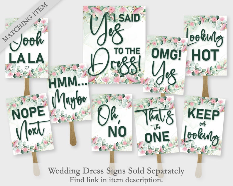 Op de afbeelding: Een verzameling borden voor trouwjurken met zinnen als "I Said Yes to the Dress!" en "Looking Hot." Elk bord heeft een witte achtergrond, donkergroene tekst en een bloemenrand met roze en groene accenten. De borden zijn bevestigd aan houten stokken.
