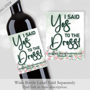 Op de afbeelding: Een wijnfles met een etiket met de tekst "I Said Yes to the Dress!" in een elegant lettertype. Het etiket heeft een aquarelontwerp met groene en roze bloemaccenten. Een bijpassende kaart wordt ook getoond.
