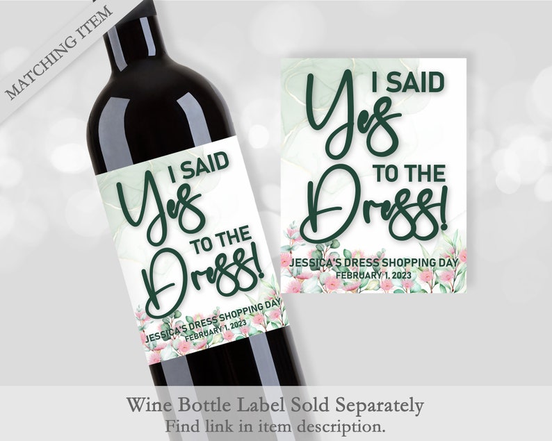 Op de afbeelding: Een wijnfles met een etiket met de tekst "I Said Yes to the Dress!" in een elegant lettertype. Het etiket heeft een aquarelontwerp met groene en roze bloemaccenten. Een bijpassende kaart wordt ook getoond.