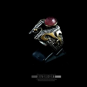 Può includere: Un anello d'argento con dettagli dorati e una grande gemma rossa e rotonda. L'anello presenta un design ornato con dettagli intricati, su sfondo nero. L'anello è un gioiello.