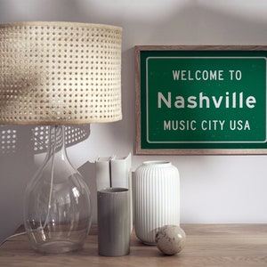 Może przedstawiać: Zielony znak w ramie z białym tekstem, kt&oacute;ry brzmi "Witamy w Nashville Music City USA". Znak znajduje się na drewnianym stole z abażurem z wikliny, przezroczystym szklanym wazonem, białym wazonem, szarym cylindrycznym wazonem i nakrapianym jajowatym przedmiotem.