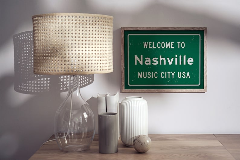 Może przedstawiać: Zielony znak w ramie z białym tekstem, kt&oacute;ry brzmi "Witamy w Nashville Music City USA". Znak znajduje się na drewnianym stole z abażurem z wikliny, przezroczystym szklanym wazonem, białym wazonem, szarym cylindrycznym wazonem i nakrapianym jajowatym przedmiotem.