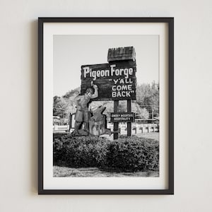 May include: Black and white photo of a vintage sign that reads "Pigeon Forge "Y'all Come Back" with a figure of a blacksmith holding a hammer and anvil. The sign is in front of a lush green bush.