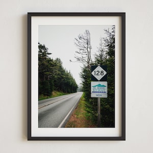 May include: A black and white sign with the number 128 on it, indicating the start of a scenic byway. The sign is on a wooden post and is surrounded by trees. A paved road leads into the distance.